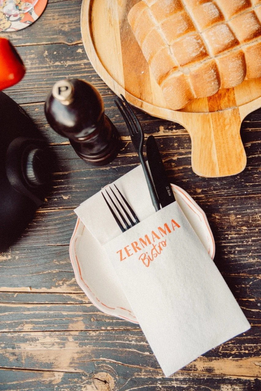 A small plate with a napkin labeled "Zermama Bistro" holding a fork and a knife, a pepper grinder, a black object, and a loaf of sliced bread on a wooden table.