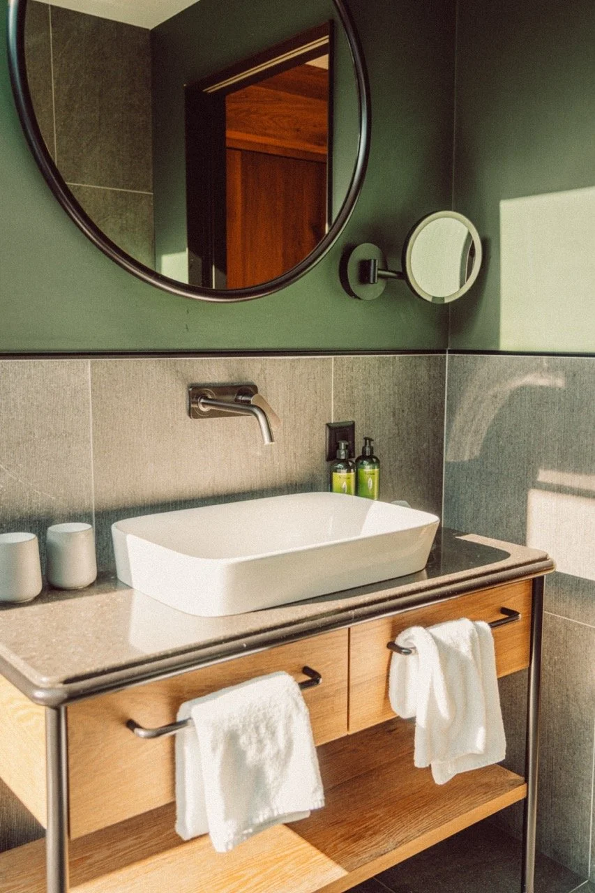 Bathroom sink with a white vessel basin, wall-mounted faucet, and a wooden cabinet with towels hanging. Above the sink, a round mirror and a smaller adjustable mirror are mounted on a green wall. Two soap or lotion bottles are on the counter, and the
