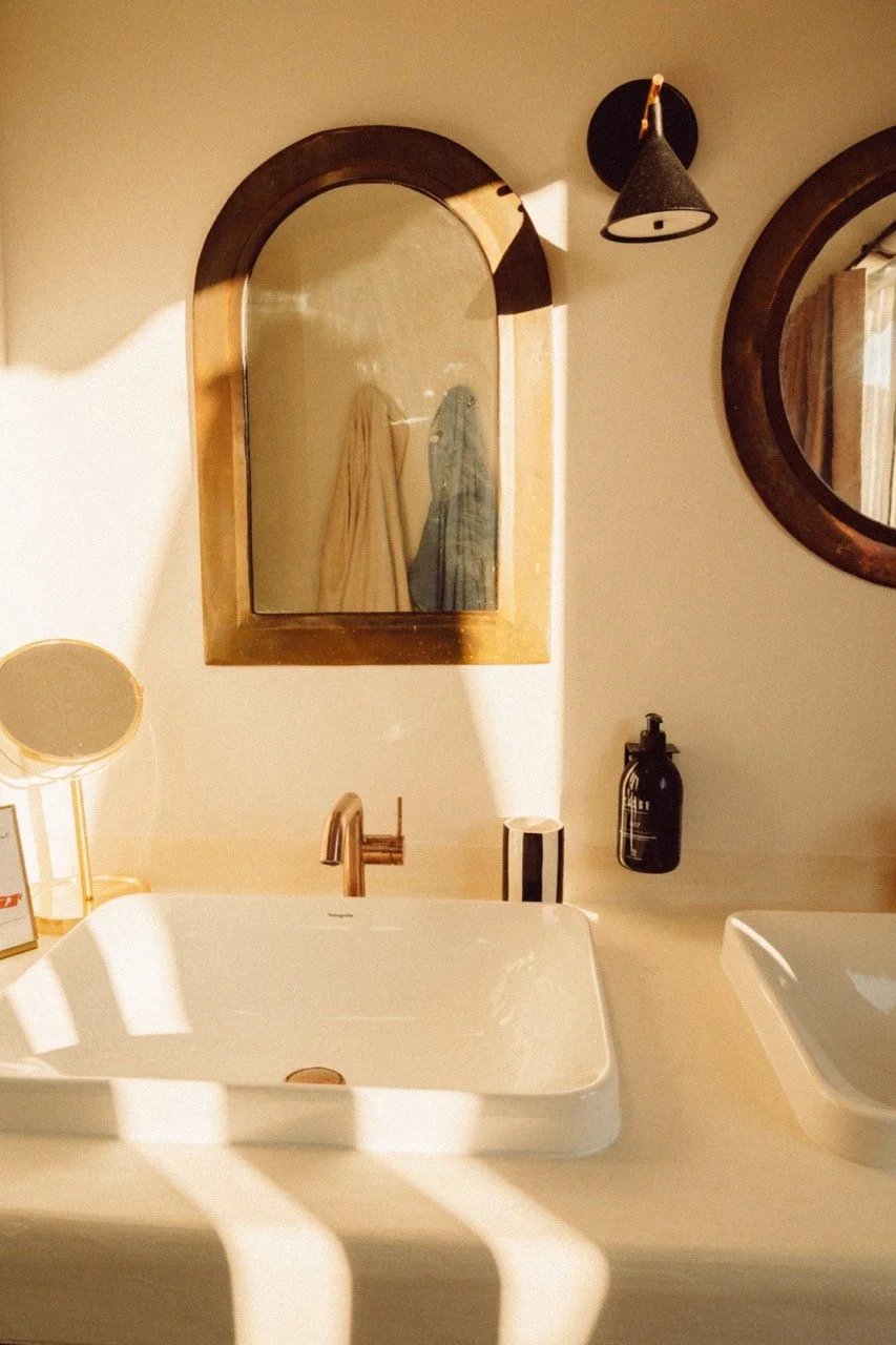 Bathroom sink area with a rectangular white vessel sink, a bronze faucet, a small round mirror, a black soap dispenser, a black wall-mounted light fixture, and an arched mirror on the wall, with sunlight casting shadows.