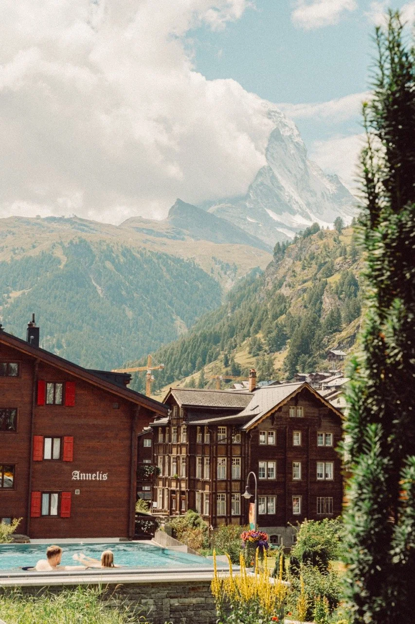 People swimming in a pool in front of a chalet-style hotel with the name Annelis, set against a backdrop of green mountains and the Matterhorn mountain in the Swiss Alps.