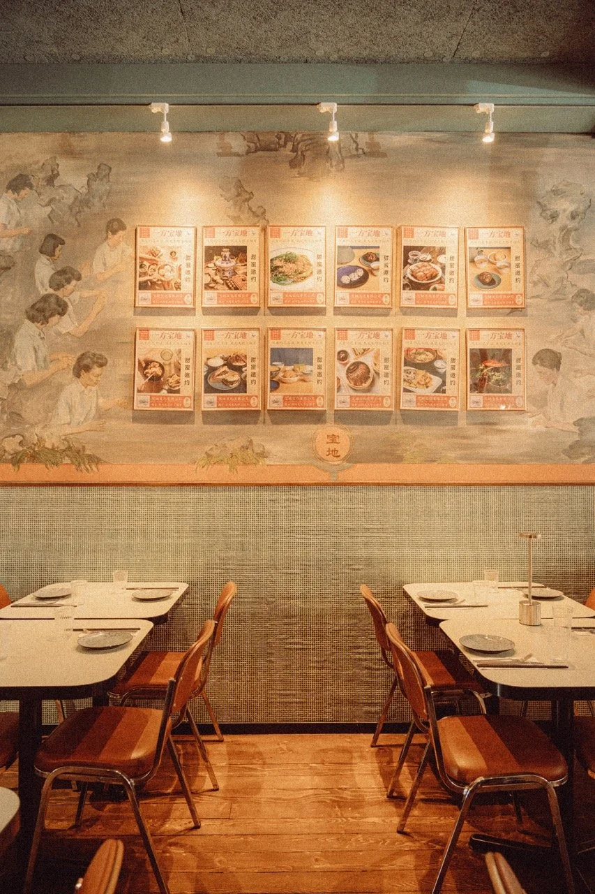 Interior of a restaurant with two white tables set with plates, cups, and utensils, and a mural on the wall advertising Asian dishes.