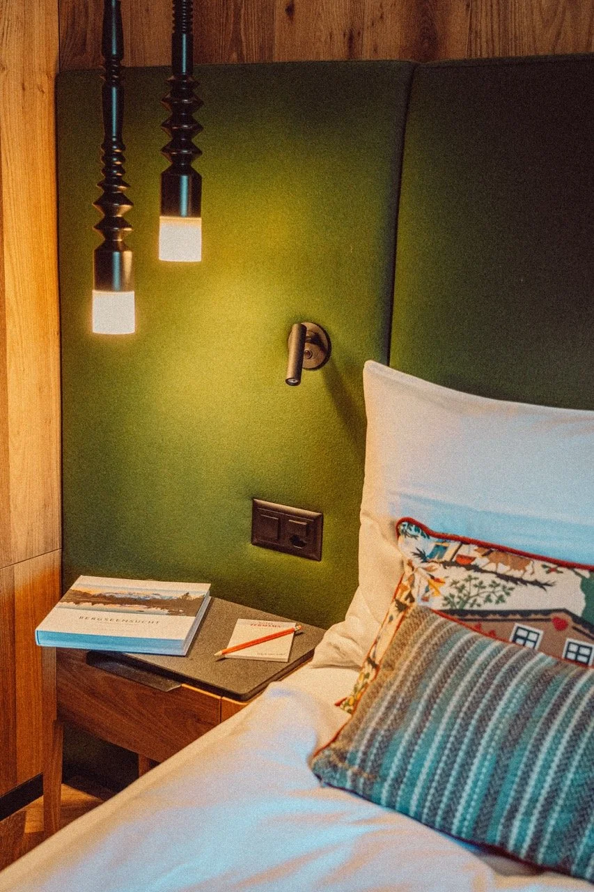 Close-up of a hotel bed with white linens, colorful decorative pillows, and a green headboard. A nightstand holds a book, a notepad, and a pen. Hanging pendant lights illuminate the area.
