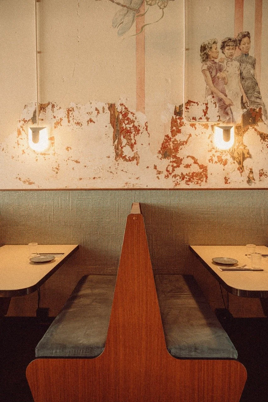 Empty restaurant booth with seating and tables set for dining, a wall with peeling paint and faded mural of three women in vintage clothing, and two modern wall-mounted light fixtures.