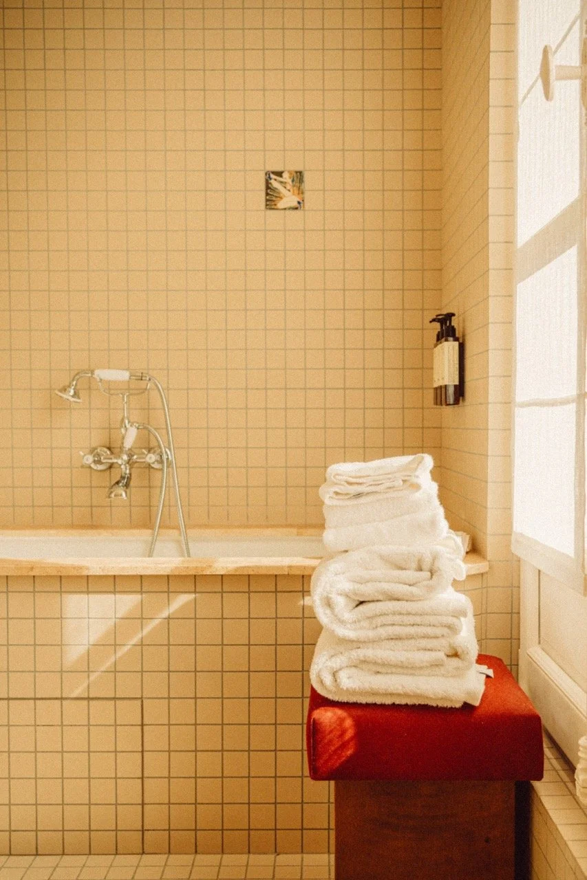 Stacked white towels on red cushioned stool in a bathroom with beige tiled walls, bathtub with faucet, and a soap dispenser on the wall.