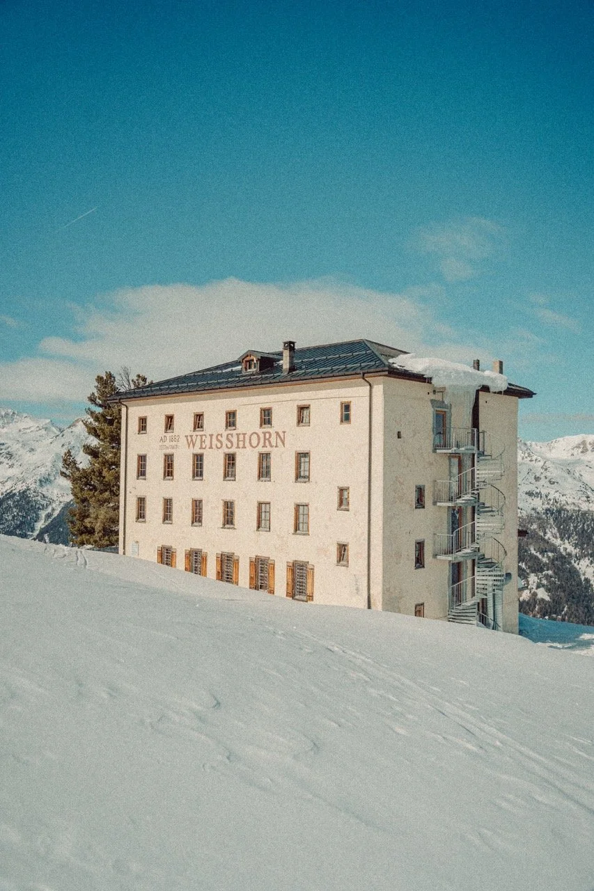 A large, historic hotel named Weisshorn, built in 1882, surrounded by snow-covered landscape and mountains, with a blue sky overhead.