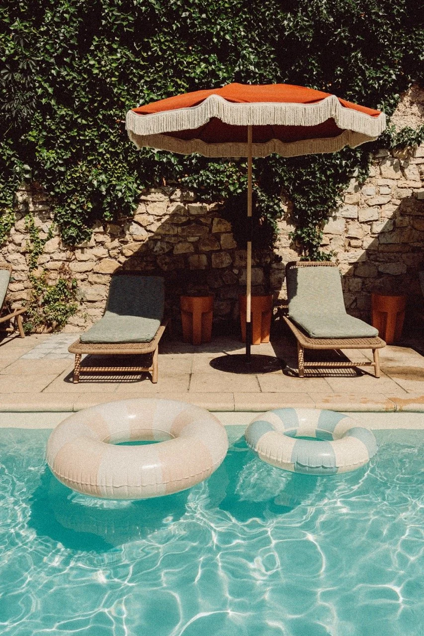 A swimming pool with two inflatable rings floating on the water, two lounge chairs with cushions, a large orange and beige umbrella providing shade, and a stone wall with greenery in the background.