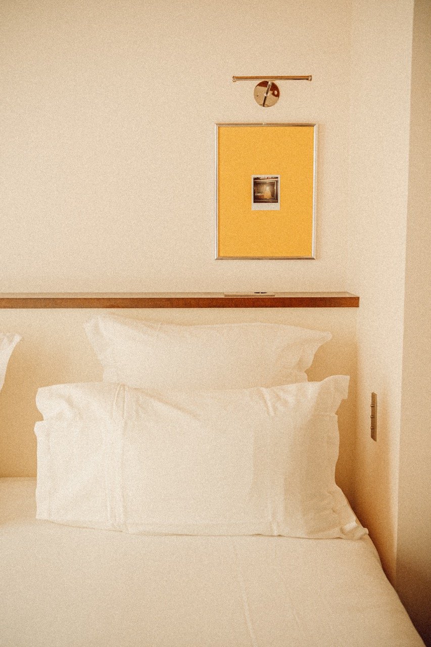 Close-up of a neatly made bed with white pillow and sheet, a yellow bulletin board on the wall above, a small picture on the bulletin board, and a wall-mounted clock.