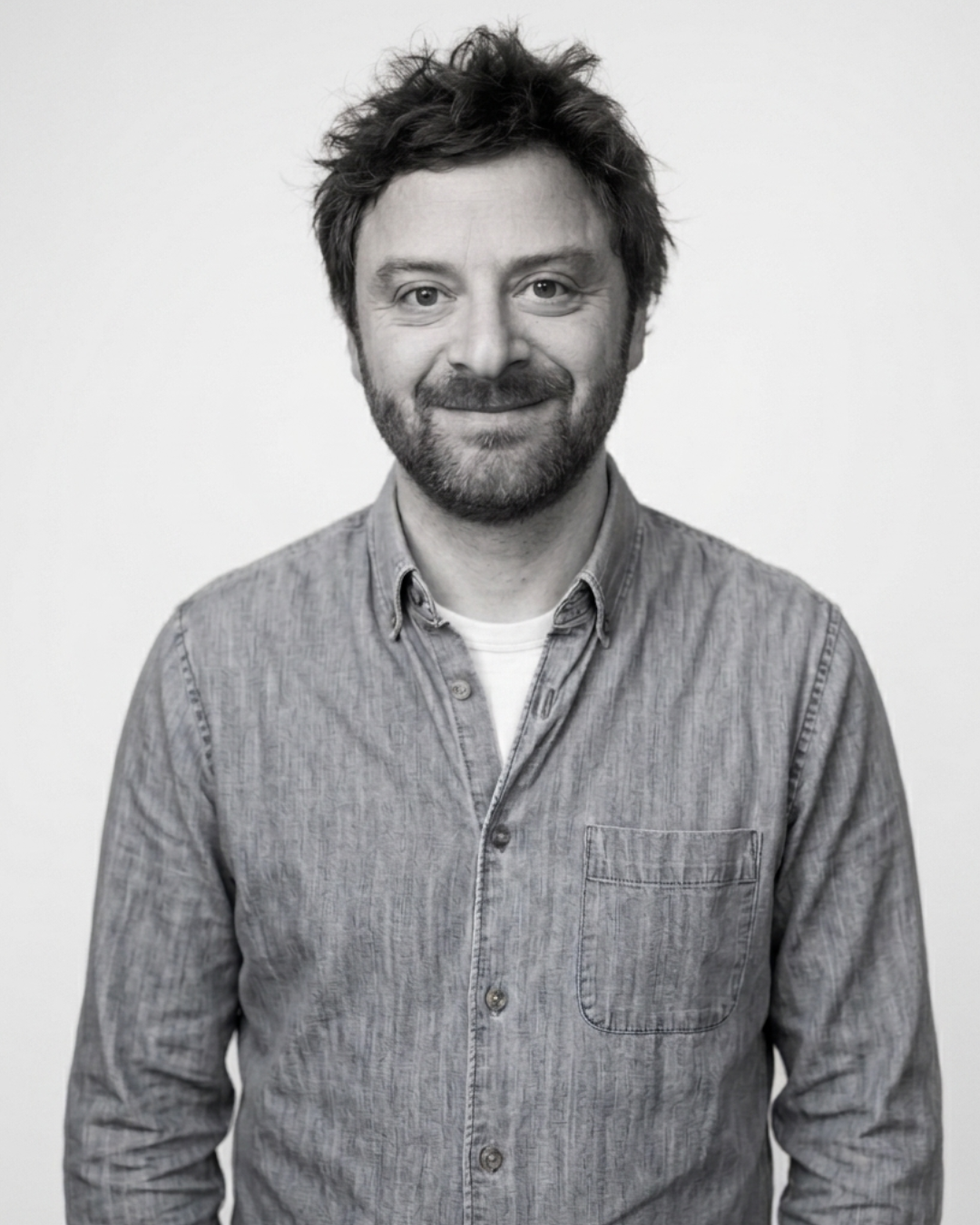 Black and white portrait of a man with messy hair and a beard, smiling, wearing a button-up shirt over a t-shirt, standing against a plain background.
