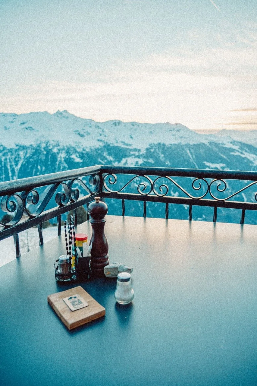 Table with salt and pepper shakers, condiments, sugar packet, rocks, and napkin holder on a balcony overlooking snow-covered mountains under a cloudy sky.