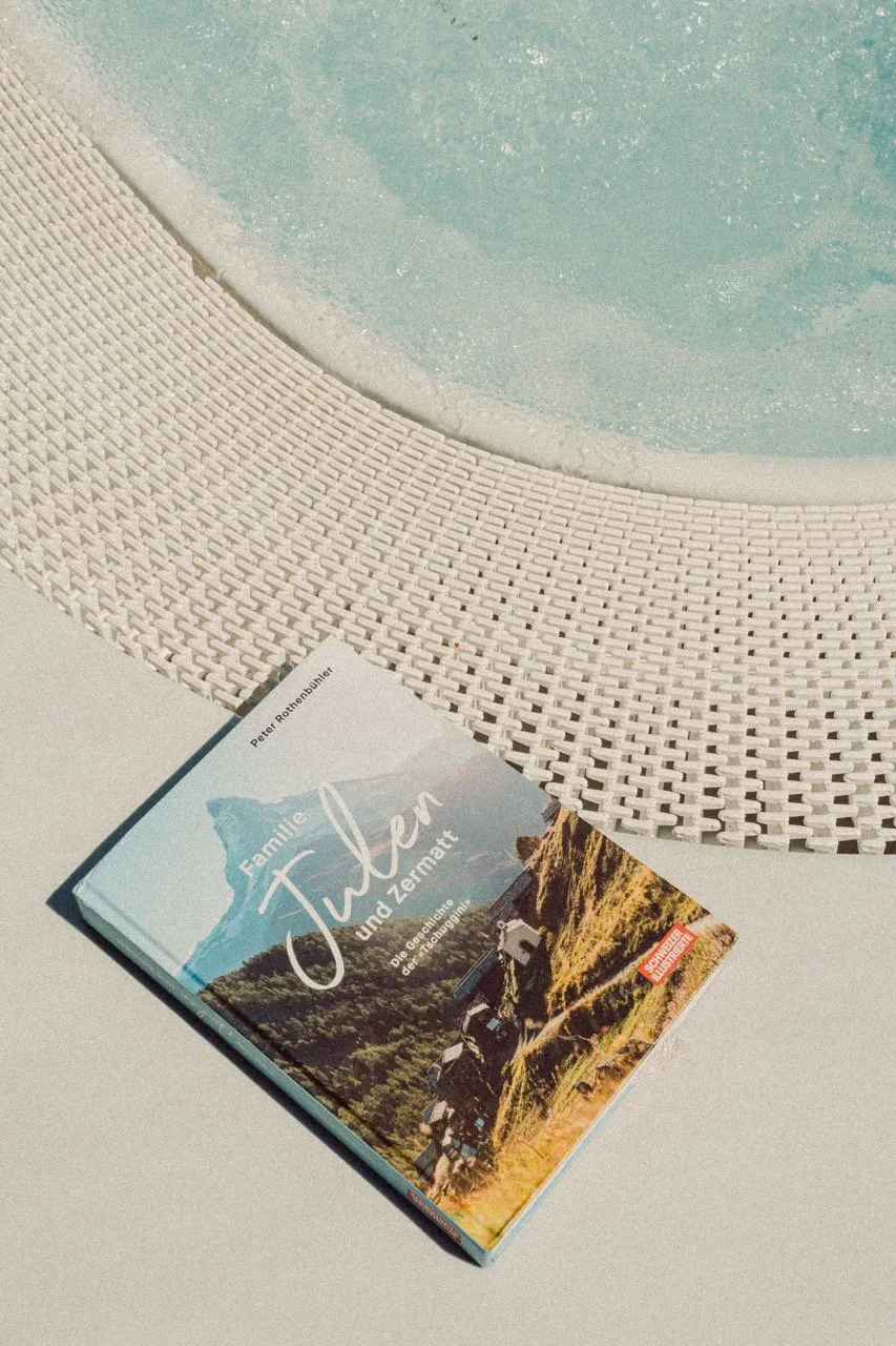 A book titled 'Familie Tüfeln und Zermat' resting on a white surface next to a textured round table with a light-colored woven pattern. The book features a landscape photo of mountains and a trail with small vehicles, and has some additional text and