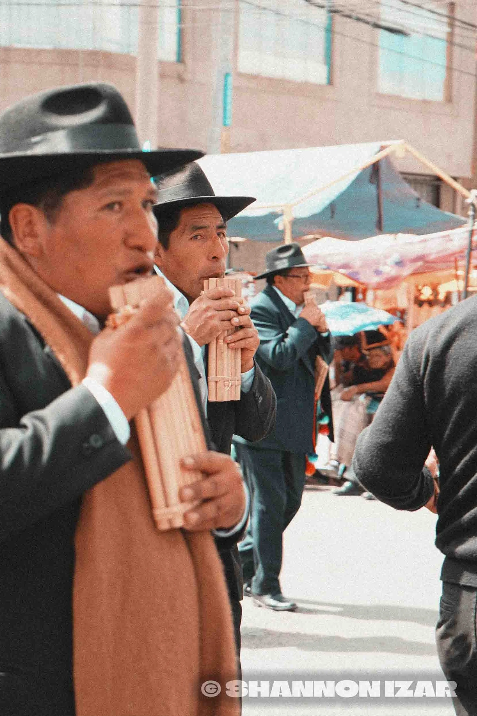Des hommes portent des sombreros noirs, certains jouent de la flute, dans une rue animée avec des étals et un parasol coloré.