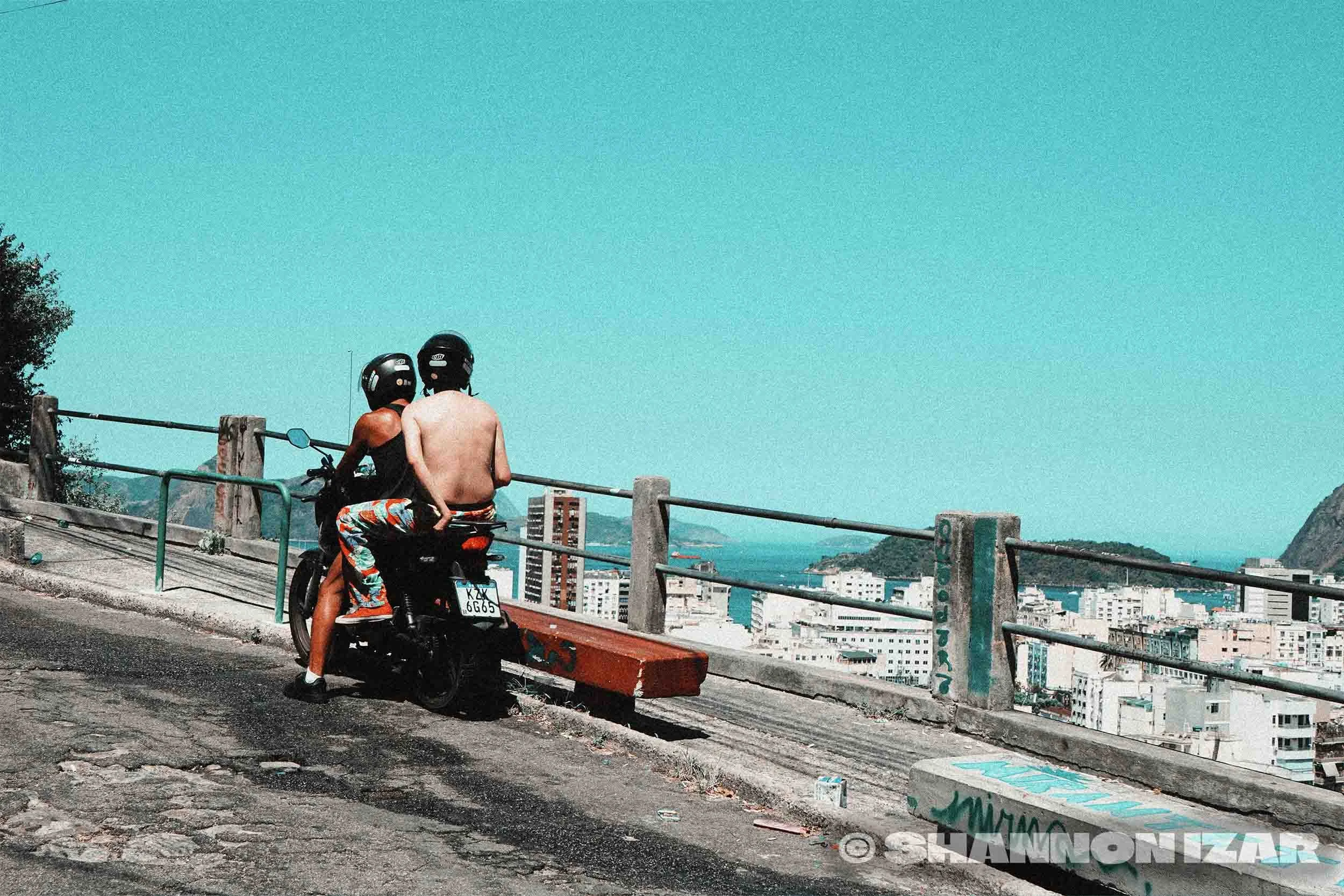 Deux hommes, dont un torse nu, portent des casques de moto et sont assis sur une moto stationnée au bord d'une route. Ils regardent un paysage urbain avec des bâtiments et une montagne au loin, sous un ciel bleu.