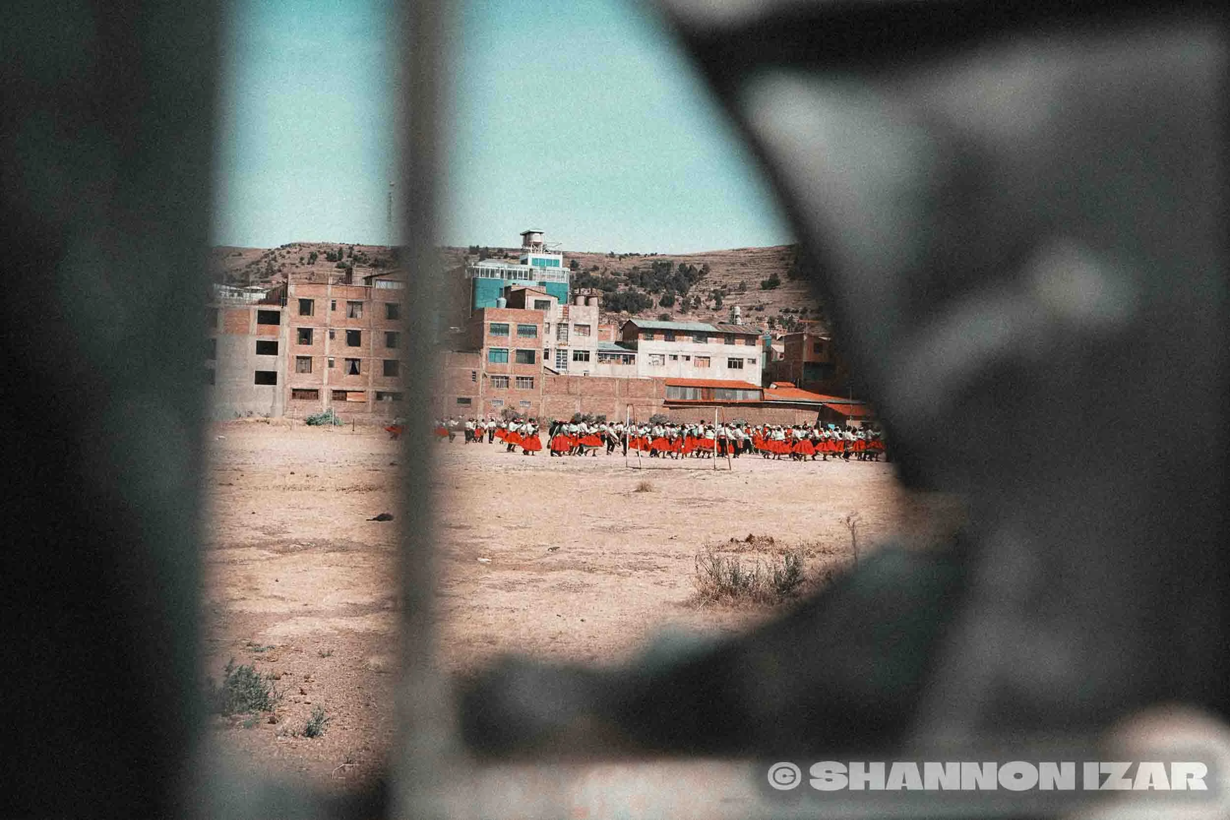 Une vue de Puno au Pérou. Vue entre des barres métalliques, avec des bâtiments incomplets et un groupe d'enfants en uniformes rouges jouant dans un terrain poussiéreux au premier plan, et des collines en arrière-plan.