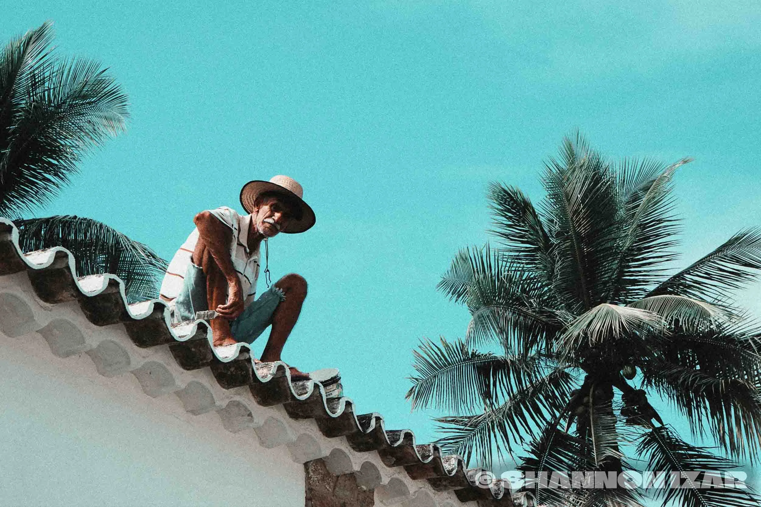 Un homme assis sur le toit d'une maison, portant un chapeau de paille, avec un ciel bleu en arrière-plan et des palmiers à proximité.