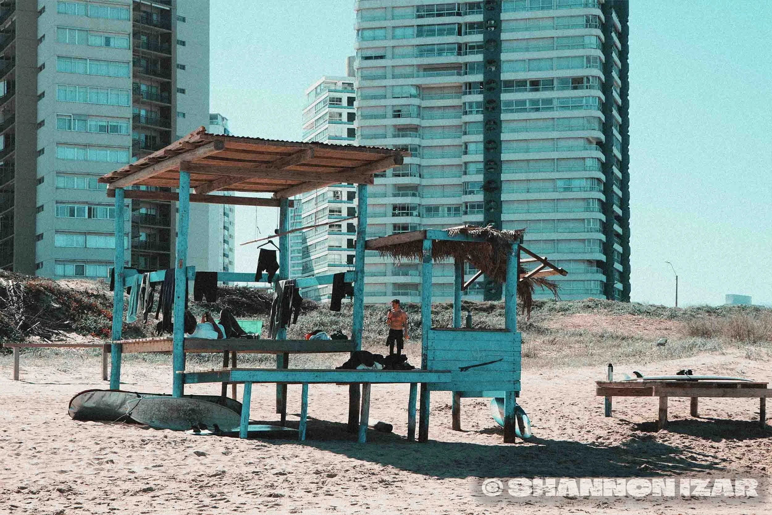 Plage urbaine avec cabanes en bois, kayak échoué, jeunes hommes, bâtiments modernes en arrière-plan.