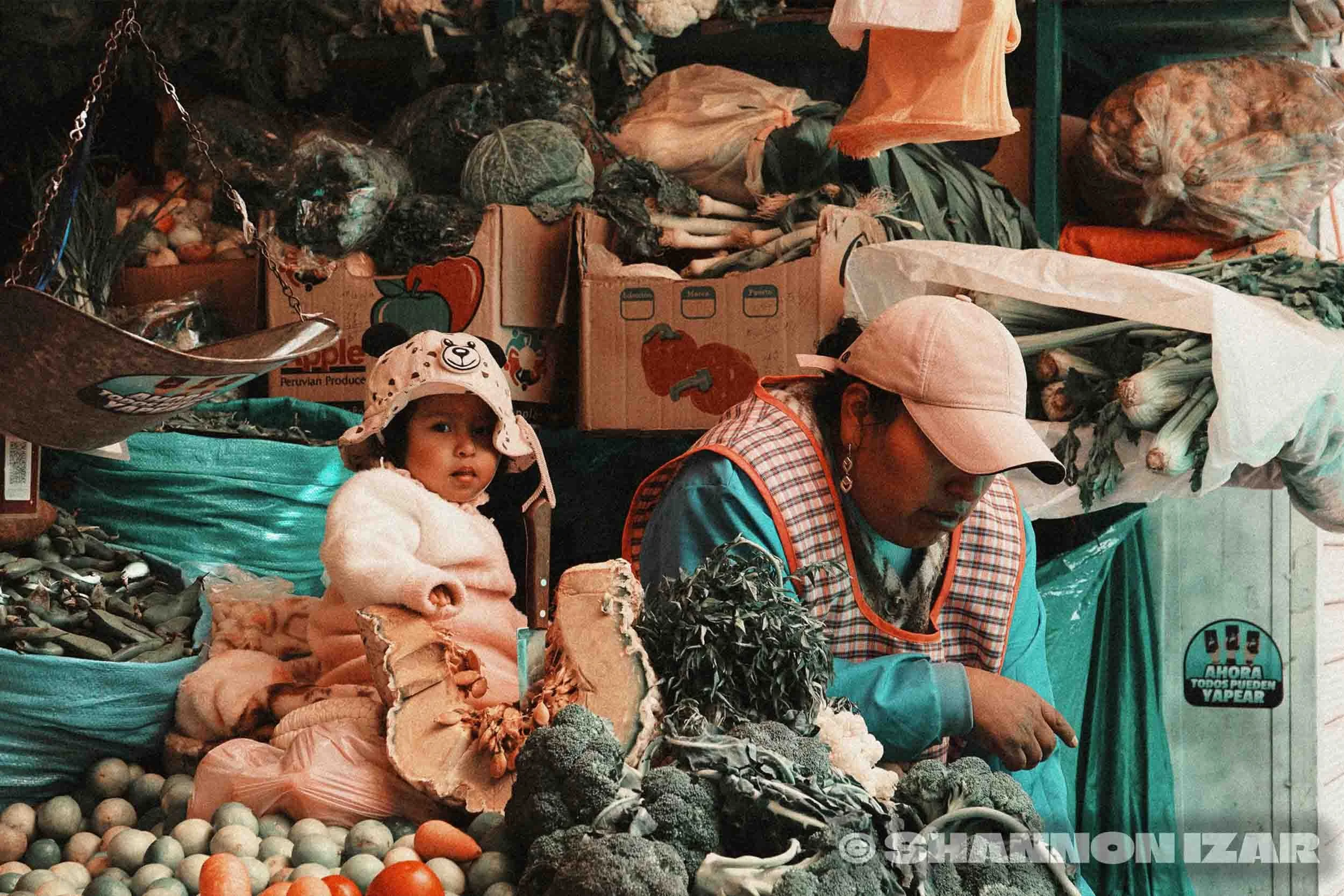 Vendeurs de marché avec des légumes variés comme brocolis, oignons et tomates, et deux enfants, l'un portant un chapeau en forme d'ours et l'autre en bonnet, en arrière-plan des boîtes de produits.