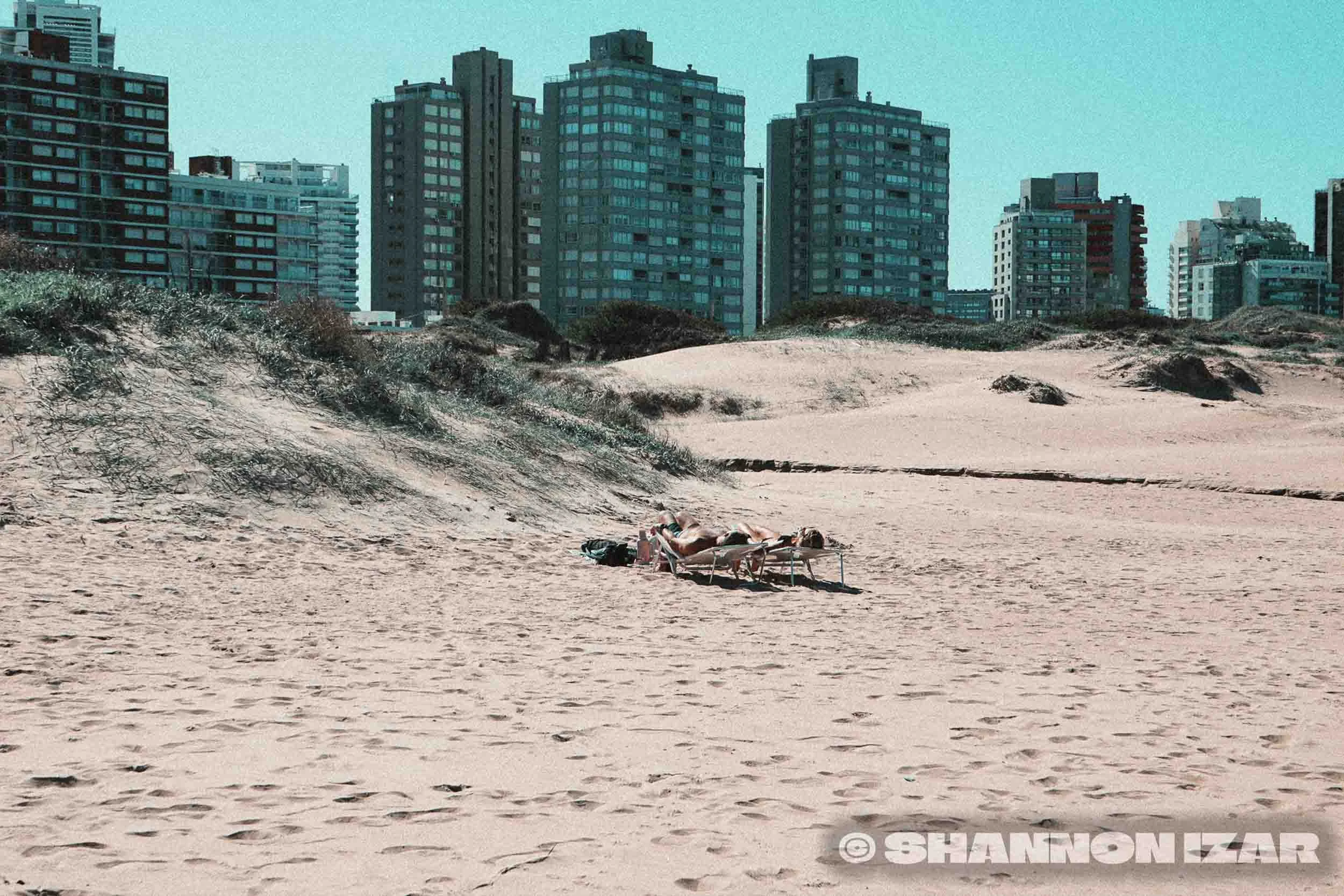 Personne allongée sur un transat sur la plage, avec un paysage de hautes tours en arrière-plan.