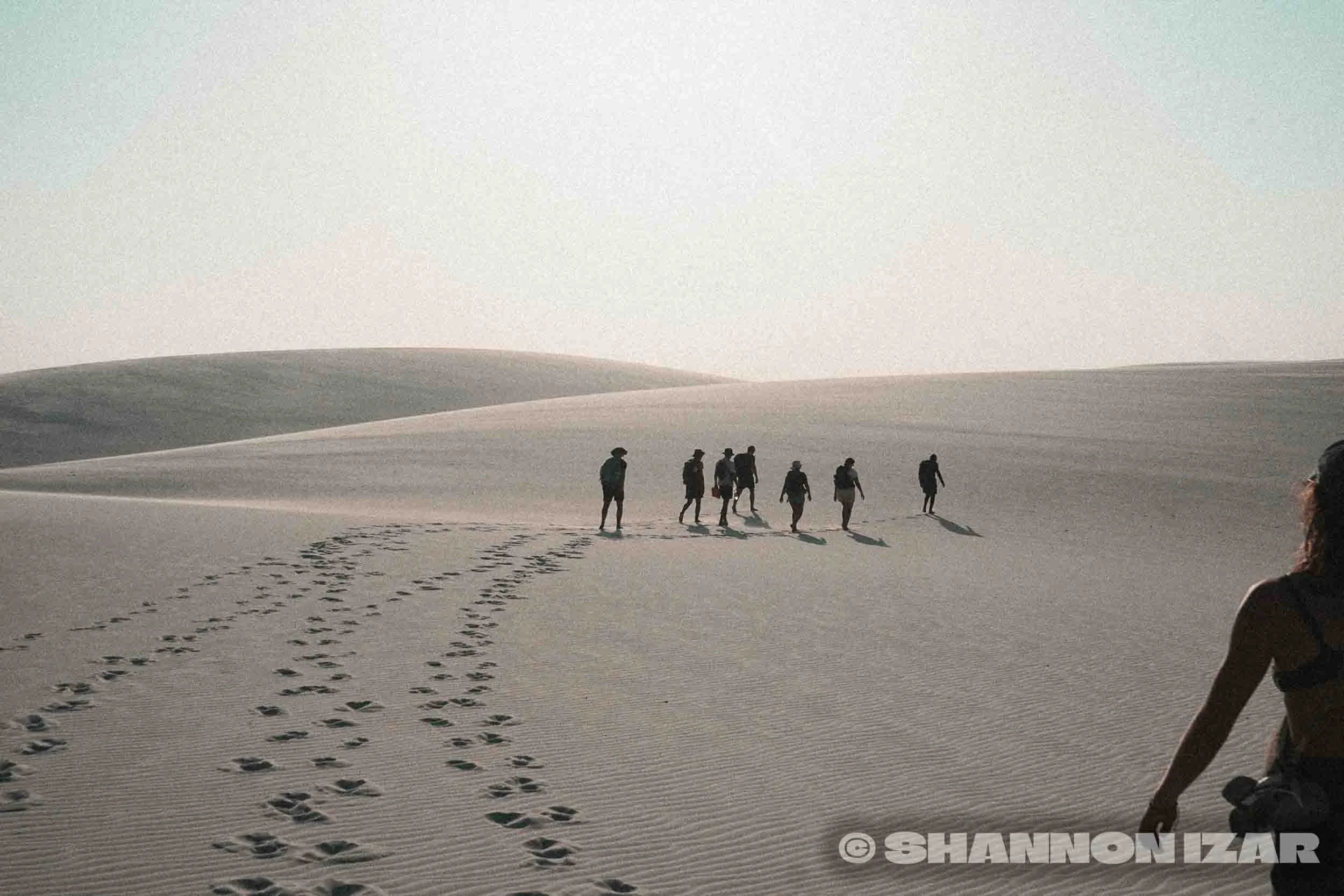 Groupe de sept personnes marchant à travers un désert de sable, laissant des empreintes derrière eux, sous un ciel clair et dégagé.