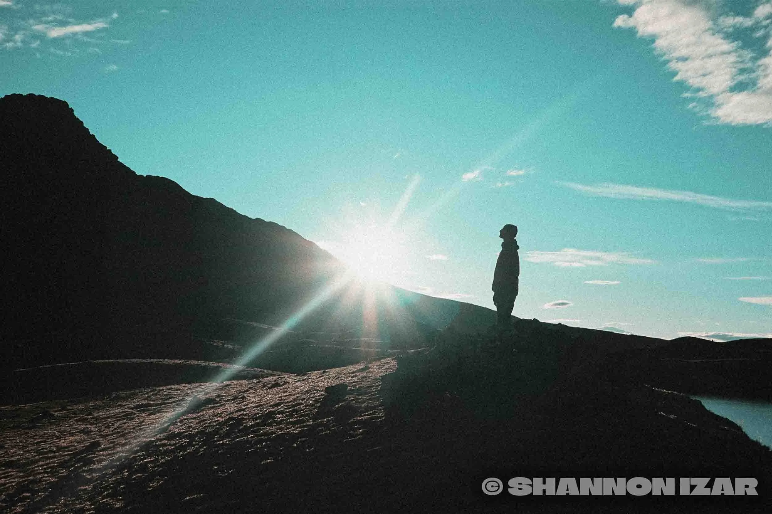 Silhouette d'une personne debout sur une roche au bord d'un lac, avec une montagne en arrière-plan sous un ciel bleu avec quelques nuages, le soleil se levant ou se couchant derrière la silhouette.