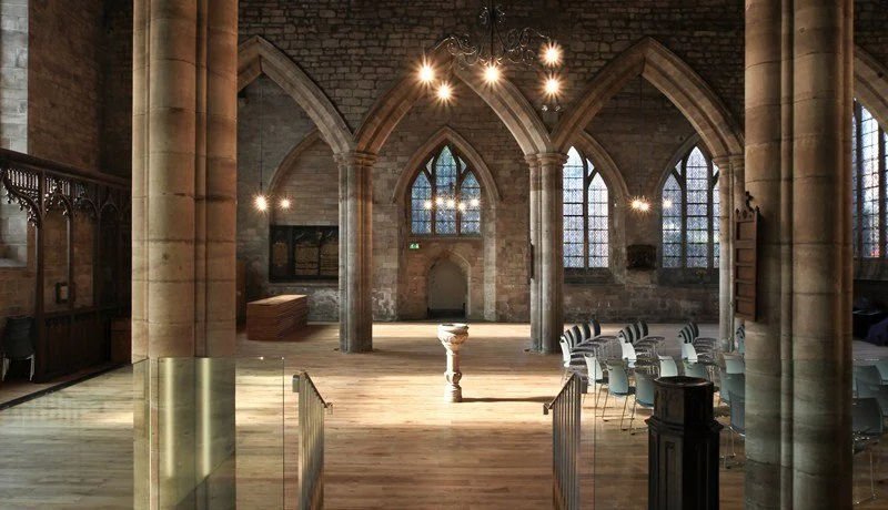 Interior of a historic church or cathedral with stone walls, arched windows, and wooden flooring, furnished with modern chairs and a small black desk.