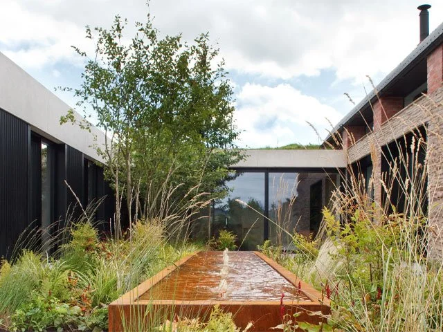 Modern outdoor courtyard with a water feature, surrounding greenery, and contemporary buildings with large windows.