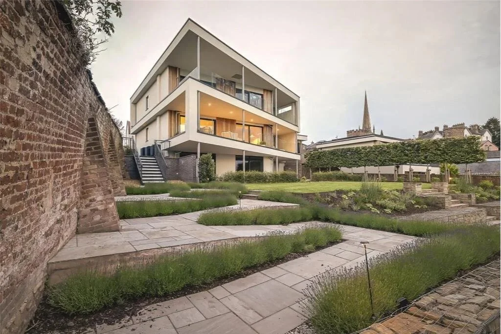Modern multi-story house with large windows and balconies, situated on a landscaped yard with paved walkways, shrubs, and a brick wall on one side.