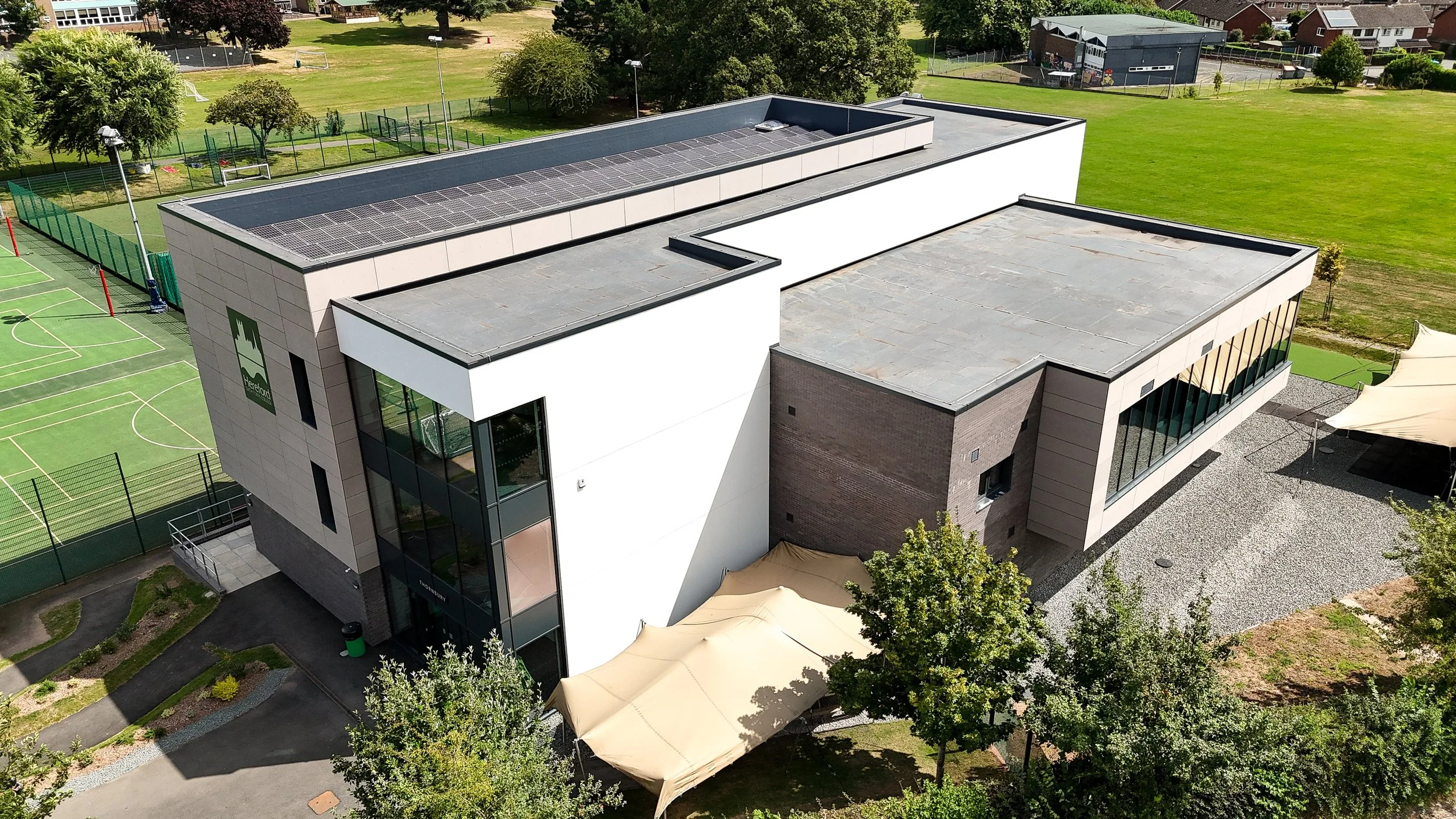 Aerial view of a modern school building with solar panels on the flat roof, surrounded by sports fields, trees, and grassy areas.