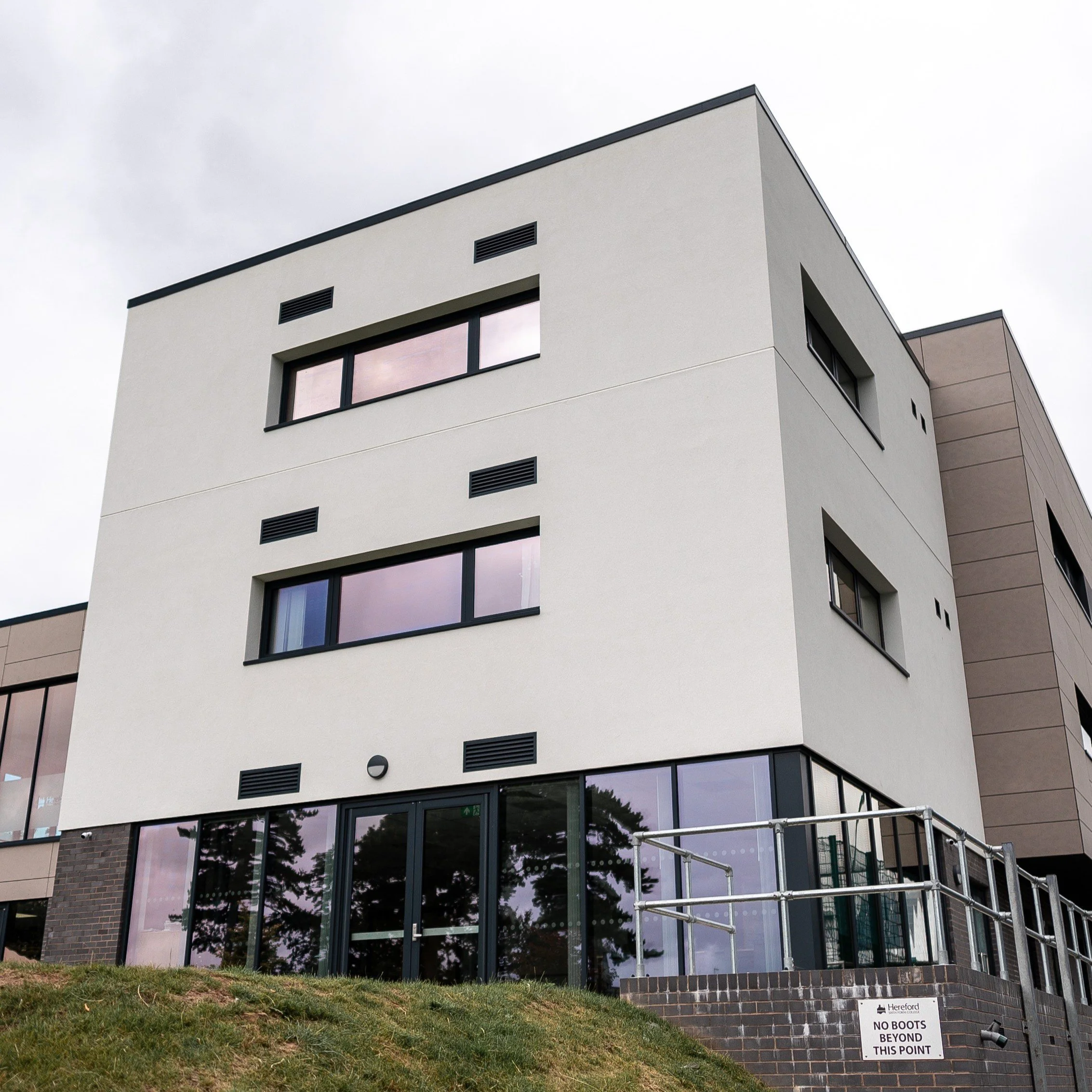 Modern multi-story building with large windows, a brick base, and scaffolding in front. There is a sign at the bottom that reads 'NO BOOTS BEYOND THIS POINT.'