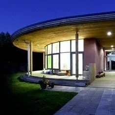 Modern house with a rounded glass exterior wall, illuminated at night, featuring a small porch and a well-manicured lawn.
