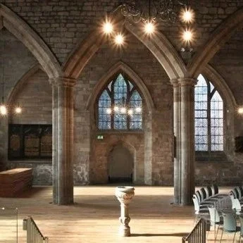 Interior of an old stone church with high arched ceilings, large stained glass windows, chandeliers, and wooden flooring.