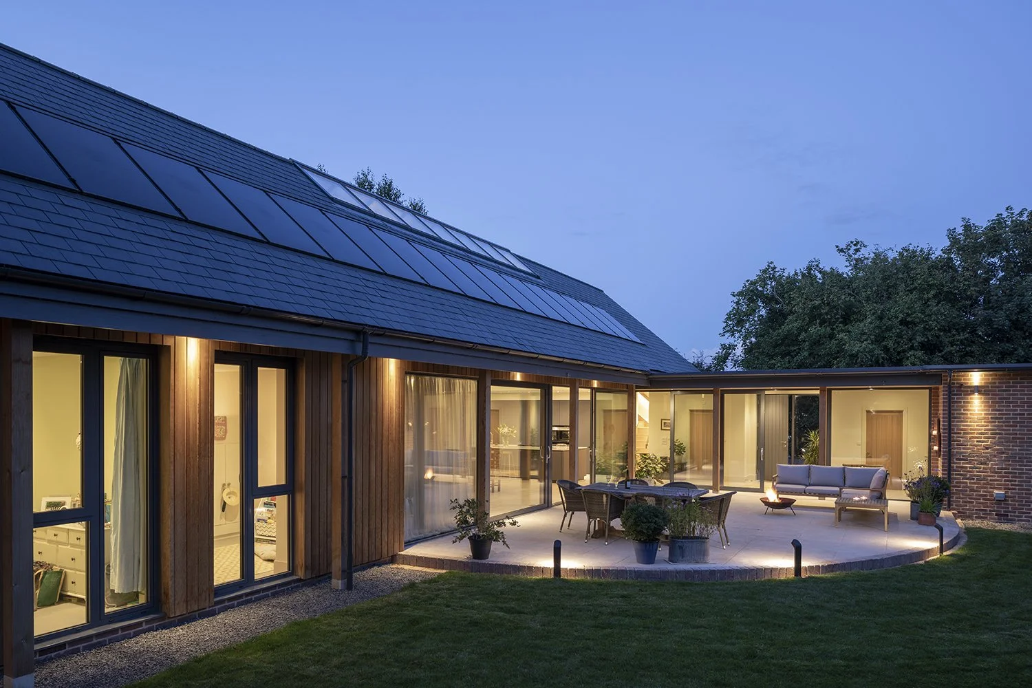 A modern house during evening with large glass windows, a curved outdoor patio with furniture, potted plants, and warm lighting, surrounded by trees and a grassy lawn.