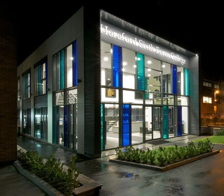 Night view of a modern two-story college building with glass walls and colorful vertical panels, illuminated with the sign 'Hereford Sixth Form College' on the front.