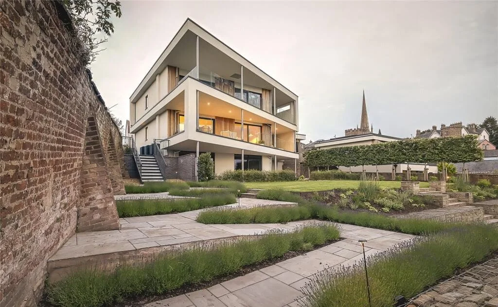 Modern multi-story house with large glass windows and balconies in a landscaped garden area, with a brick wall on one side and a church spire in the background.