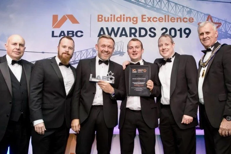 Group of six men in tuxedos standing on stage at the 2019 Building Excellence Awards, holding a plaque and LABC award, with a backdrop displaying the event name and logo.