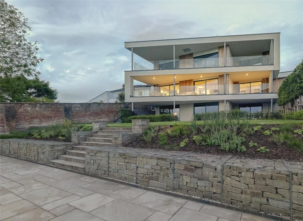 A modern multi-story house with large glass windows and balconies, situated on a raised landscaped yard with stone steps, grass, and plants, during twilight.