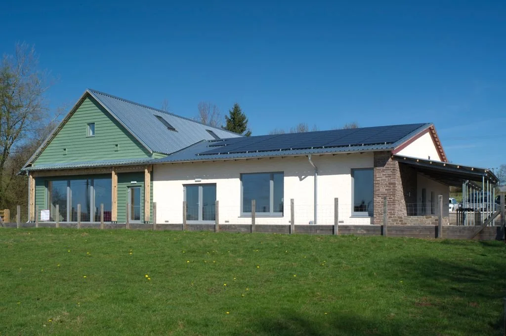 A modern house with a green and white exterior, large windows, and solar panels on the roof, surrounded by a grassy yard under a clear blue sky.