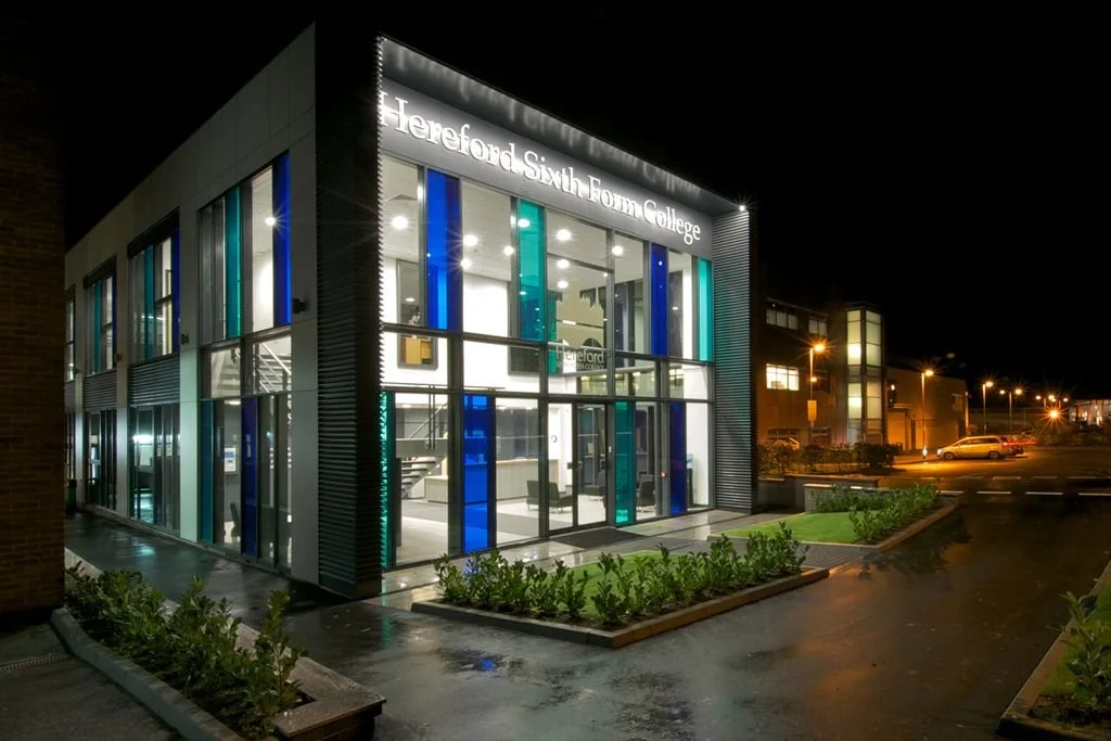 Night view of a modern educational building with illuminated sign reading 'Hereford Sixth Form College', large glass windows, and colorful vertical glass panels, surrounded by small green bushes and streetlights.