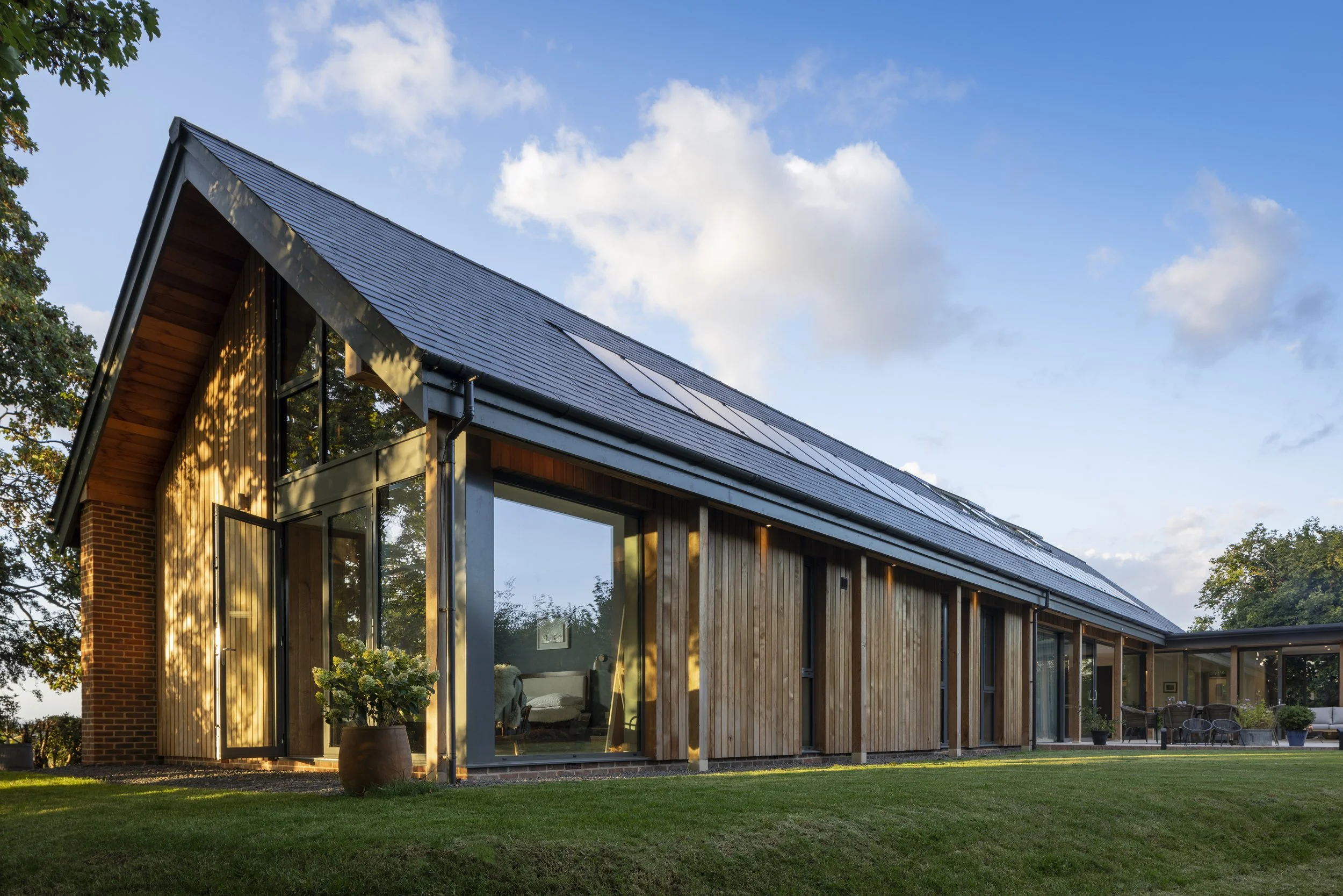 Modern house with a sloped roof covered in solar panels, large glass windows, and a well-maintained lawn under a blue sky with scattered clouds.
