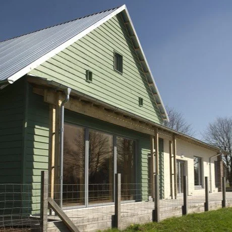 Part of a house with green siding and large sliding glass doors, with a sloped roof and a small deck, in a suburban setting.