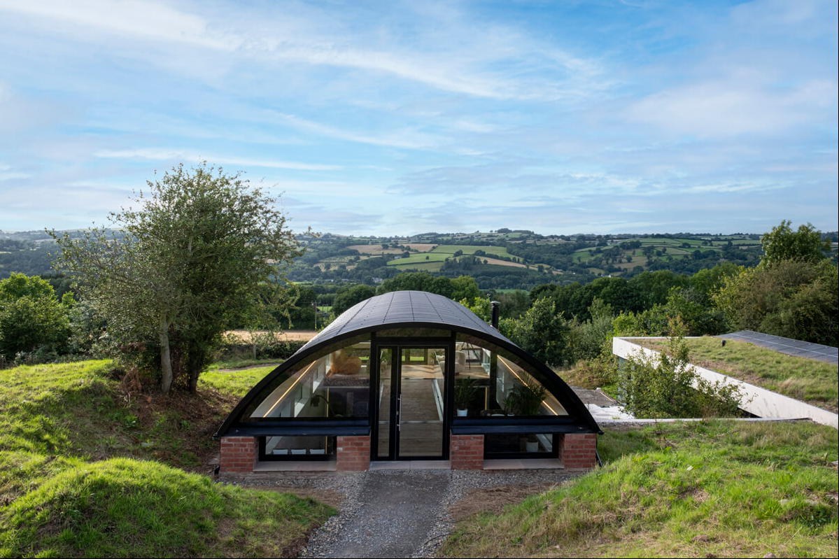 A modern, arched glass and brick house in a lush green landscape with rolling hills in the background, under a partly cloudy sky.