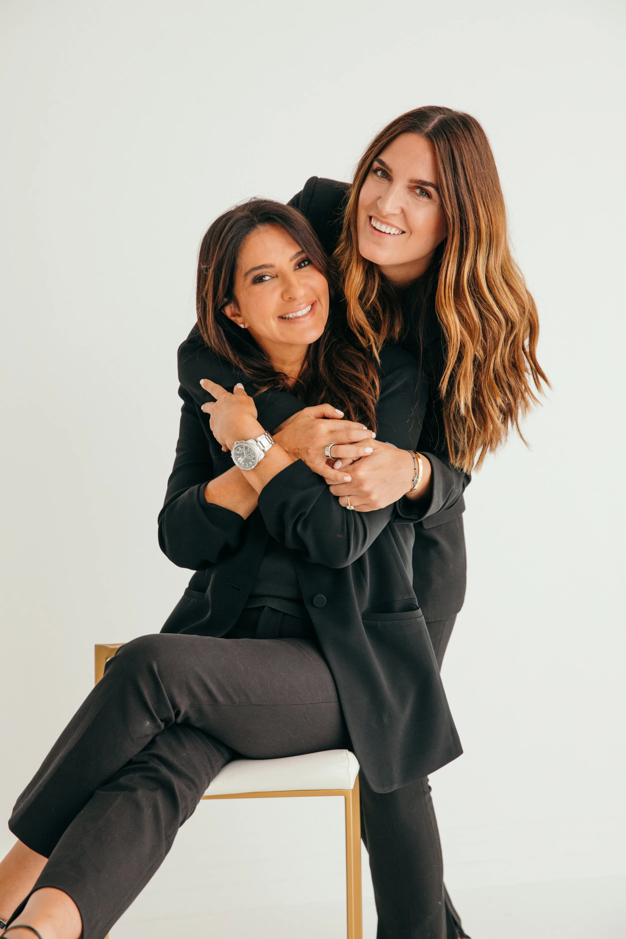 Two women smiling, one sitting on a chair with one leg over the other, the other standing behind, leaning in. They are dressed in black business attire against a plain light background.