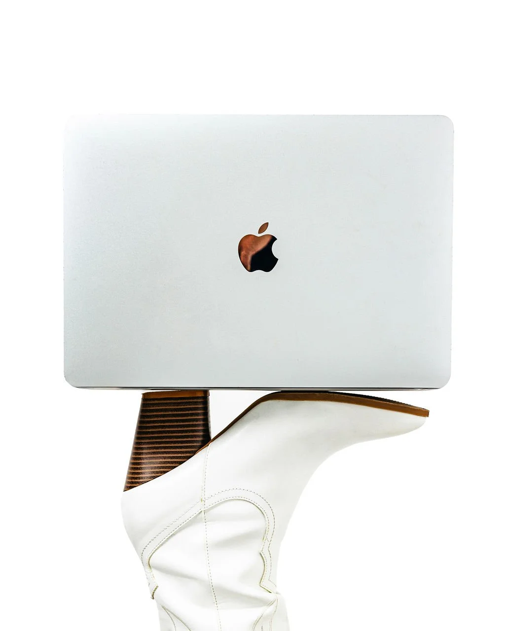 A closed Apple MacBook laptop is balanced on a white shoe with a brown heel, against a plain white background.