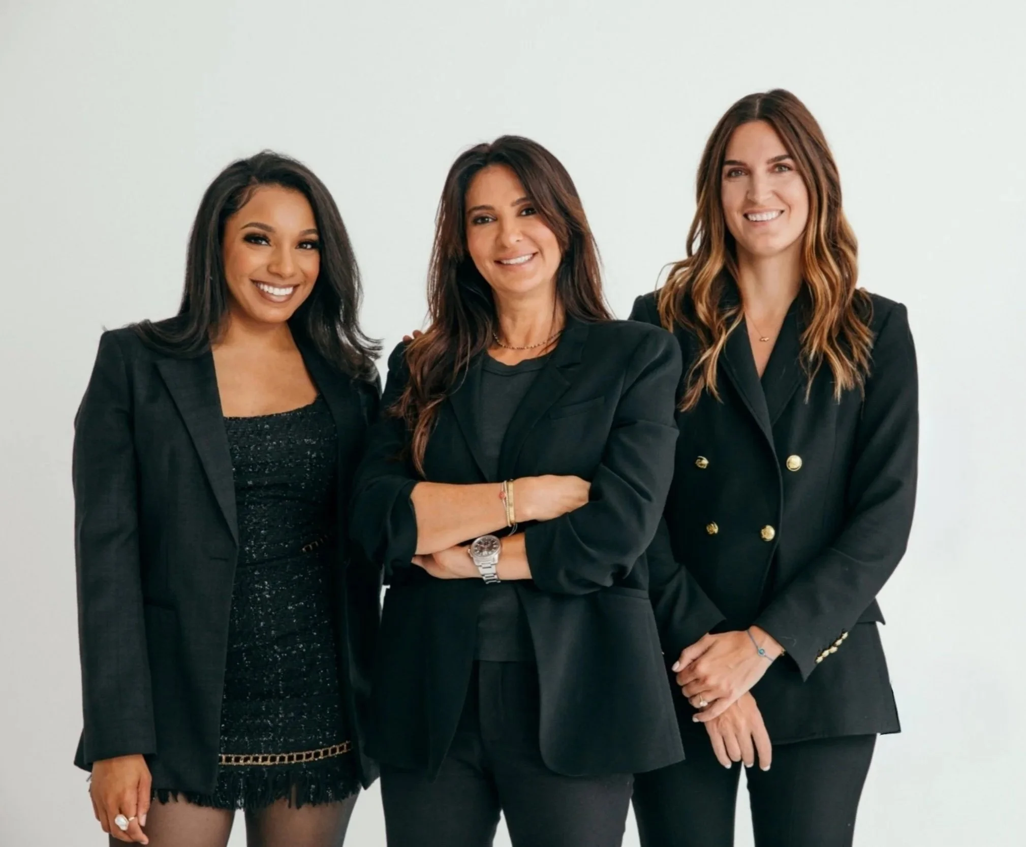 Three women dressed in black blazers standing together in front of a plain white background, smiling