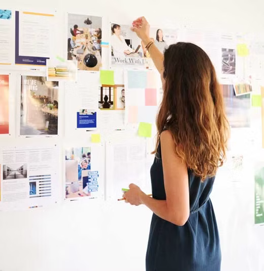 A woman with long, wavy hair in a dark dress stickers a notice on a white wall filled with various papers, photos, and sticky notes.