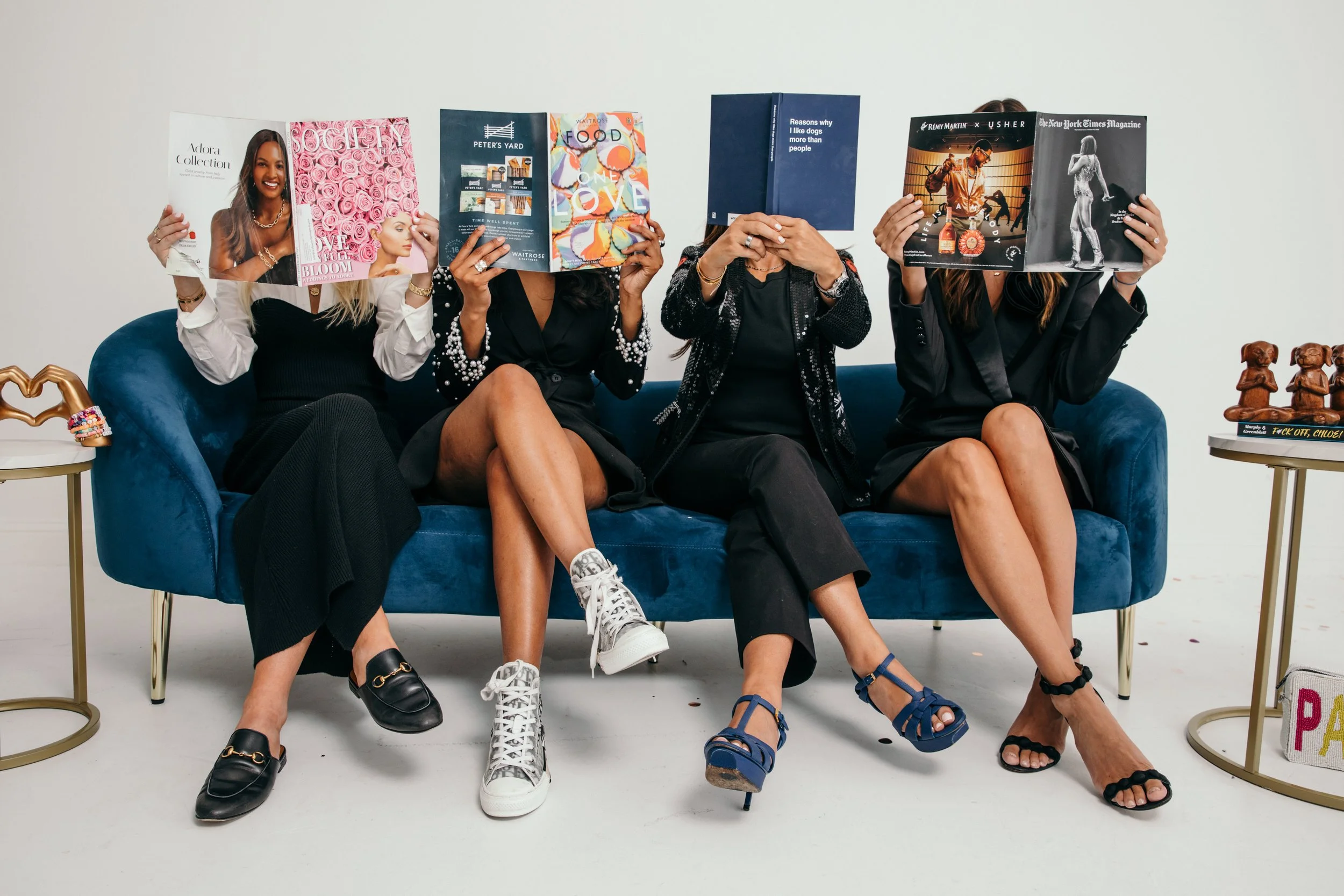 Four women sitting on a blue sofa, each holding a magazine that covers their face. The magazines feature various colorful images and text. There are two side tables with decorations, including a heart-shaped figurine and small dog sculptures. The women are dressed in stylish black and white outfits with accessories, and they are wearing different types of shoes.