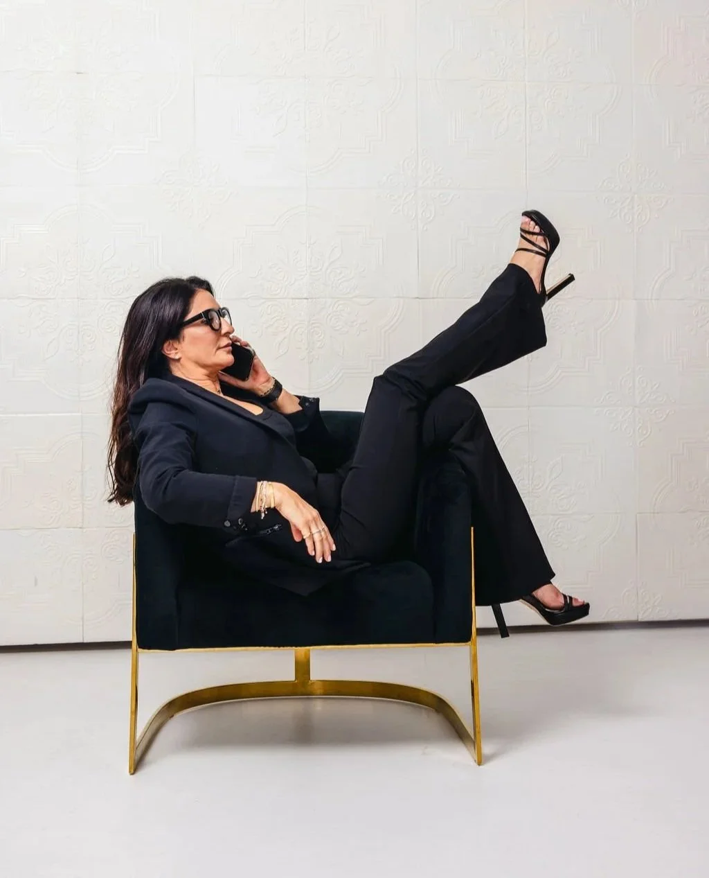 A woman with dark hair and glasses wearing a black suit, sitting on a black and gold chair, talking on a cell phone, with one leg raised and foot in a black high heel.