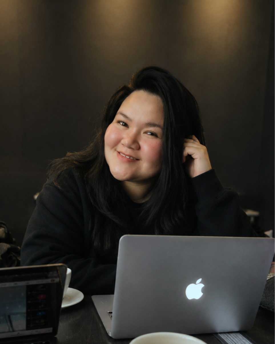 A woman with long black hair smiling at the camera, sitting at a table with a silver MacBook, a cup, and a smartphone, in a dark-colored room.