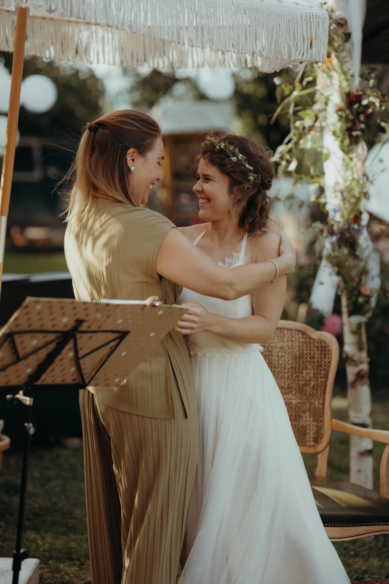 Zwei Frauen umarmen sich bei einer Hochzeit im Freien, mit Blumen und einem Sonnenschirm im Hintergrund.