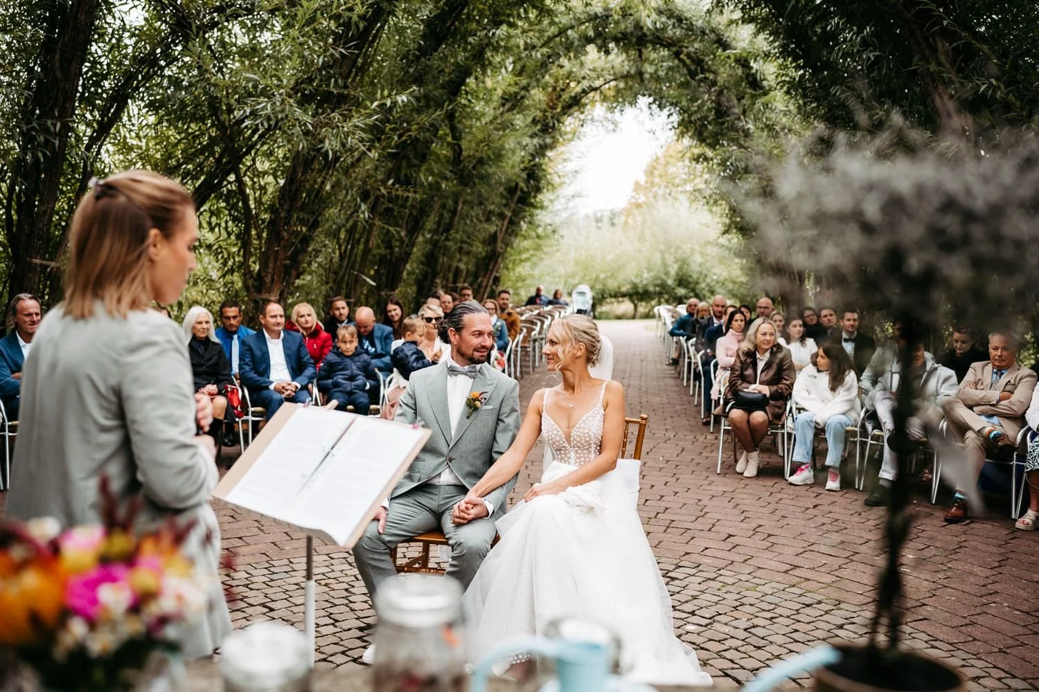 Hochzeit im Freien mit Braut und Bräutigam, die sich die Hände halten, während sie einem Traurteil lauschen, umgeben von Gästen unter Bäumen