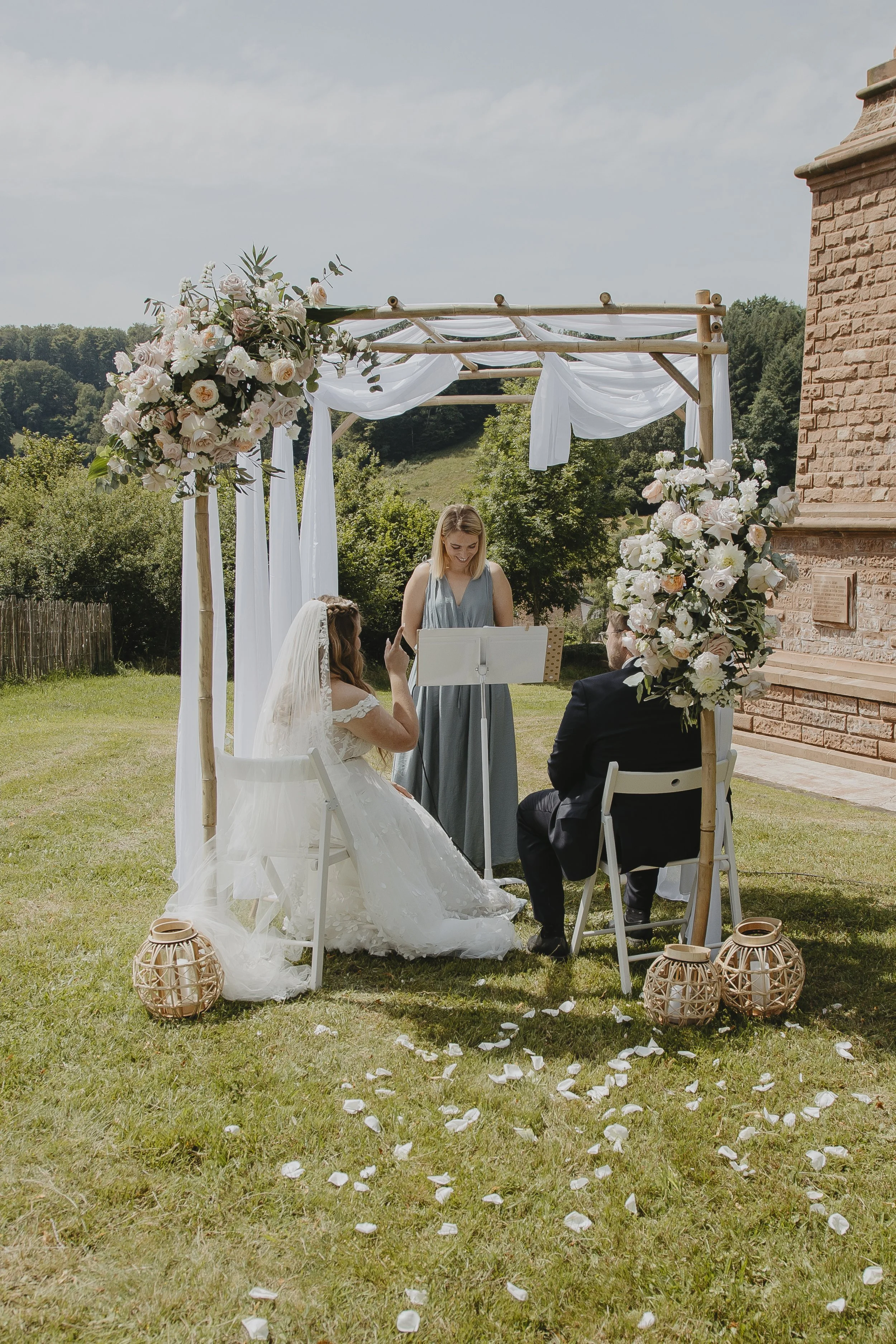 Hochzeit im Freien mit Braut, Bräutigam und Standesbeamtem unter einem Blumenbogen, auf einem Rasen mit fliegenden Rosenblättern, umgeben von Bäumen und einem roten Backstein-Gebäude.